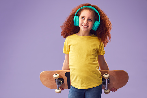 Studio Shot Of Young Girl Wearing Headphones Holding Skateboard Against Purple Background