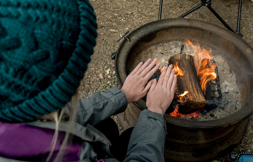 Camping lifestyle. Girl wearing beanie hat wariming hands over the campfire on a camping site. Selective focus