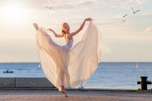 dancing ballerina in a white flying skirt and tights on the ocean embankment or on the sea beach, surrounded by seagulls in the sky.