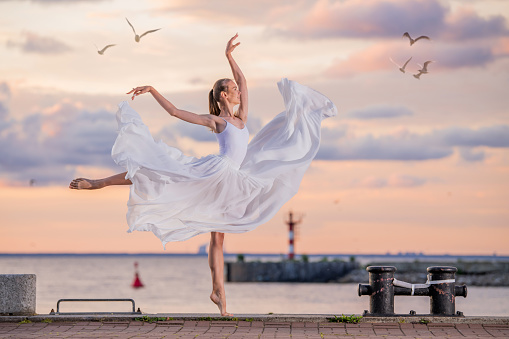 dancing ballerina in a white flying skirt and tights on ocean embankment or on the sea beach against backdrop of sunset sky