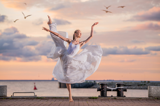 dancing ballerina in a white flying skirt and tights on ocean embankment or on the sea beach against backdrop of sunset sky