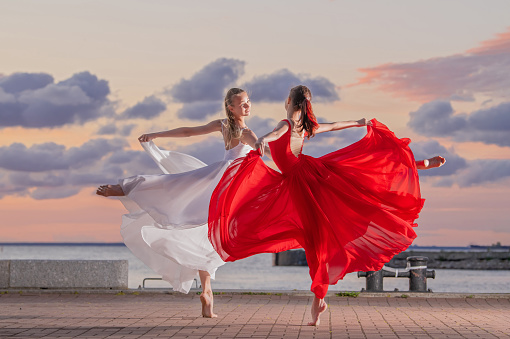 Two ballerinas in a white and red flying skirt and leotard dancing in a duet on the embankment of sea against backdrop of sunset sky