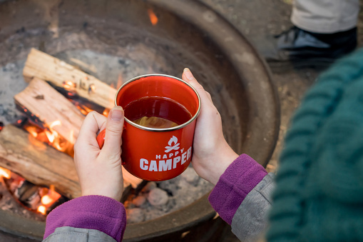 Camping lifestyle. Girl wearing beanie hat holding red enamel mug near a campfire. Happy camper