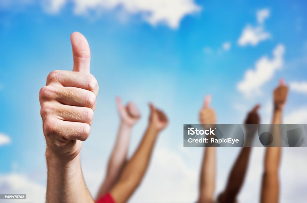 Group of people give the thumbs-up gesture of approval or permission against a beautiful summer sky outdoors Diverse group of people with arms raised, all giving the thumbs-up sign. Blue Stock Photo