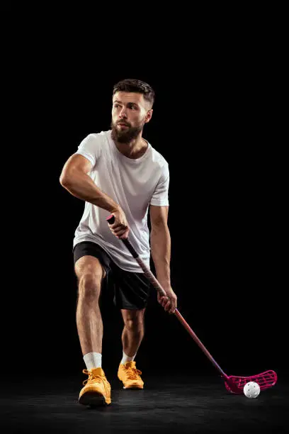 Pass. Studio shot of young man in sports uniform and sneakers playing floorball isolated on dark background. Sport, action and motion, movement, competition. Training with floorball stick and ball