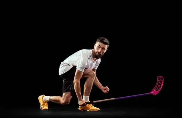 Studio shot of young man wearing sports uniform and sneakers playing floorball isolated on dark background. Sport, action and motion, movement, competition. Training with floorball stick and ball