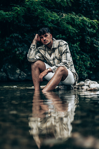 Portrait of a man in chequered shirt sitting on the rocks in the river and looking at the camera