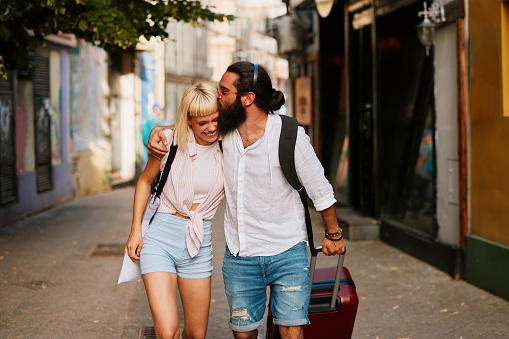 Happy young tourist couple walking through european city with suitcase