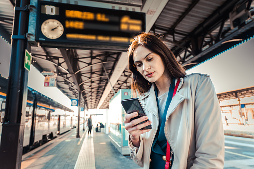 Young woman with departure times behind her waiting for her train while holding her mobile phone - Woman taking a selfie - Transportation and urban life concept