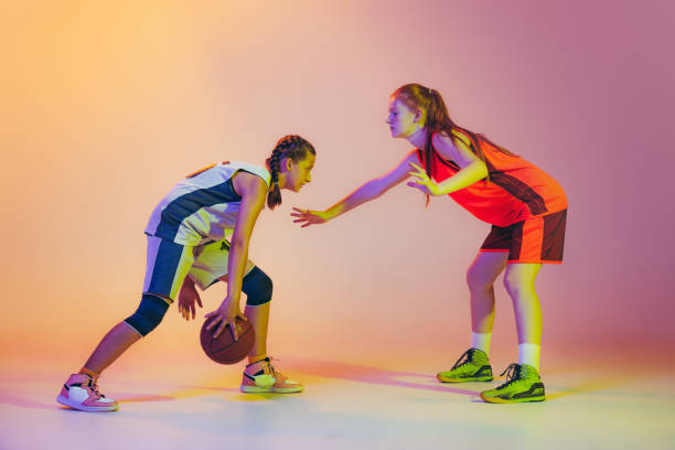 攻撃と防御。女性バスケットボール選手、若い女の子、ネオンドの背景に隔離されたバスケットボールボールで行動中の十代の若者。スポーツ、チーム、エネグリー、競争、スキルの概念 - basketball team sports healthy lifestyle isolated objects ストックフォトと画像