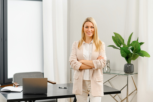 Smiling caucasian woman looking at the camera while posing arms crossed. Blonde haired lady standing at her modern office
