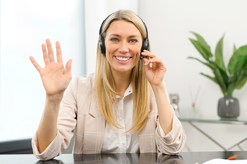 Headshot of smiling young female employee wearing wireless headset, looking at the camera, Cheerful woman in headphones taking call from colleagues or clients online, working remotely from home office