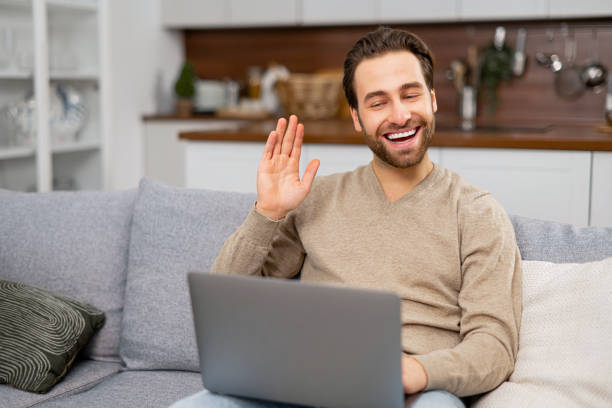 istock Cheerful carefree guy using laptop computer for video connection, looking at the webcam and waving hand. Man sitting at the sofa and making video call 1409491523