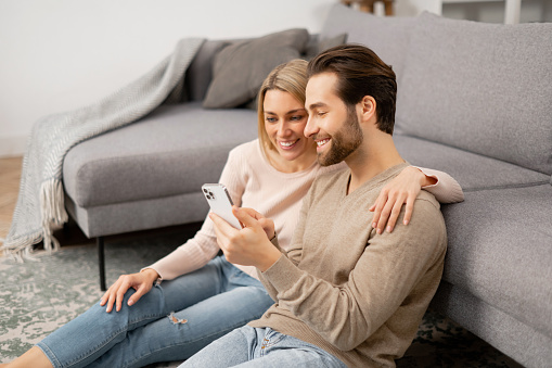 Caucasian couple with smartphone spending leisure time in social networks, scrolling feed, watching videos. Woman sitting at the floor near her boyfriend