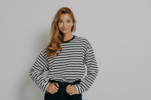 Young confident charming woman in striped blouse and casual jeans standing with hands on waist, looking at camera with approving smile, isolated over grey studio wall with positive face expression