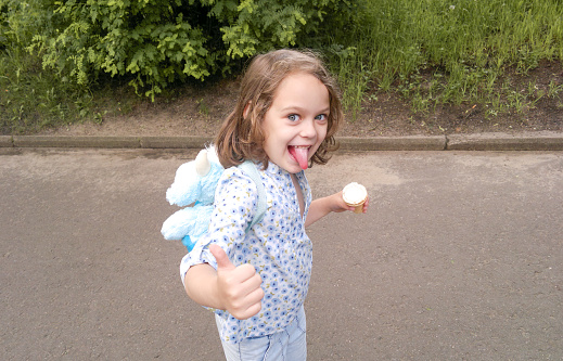 Funny blond blue eyed girl with ice cream stuck out tongue, shows Like gesture. Naughty playful.