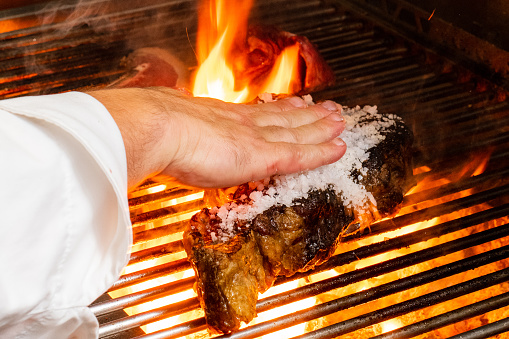 Cook's hand distributes coarse salt in veal steak on the grill