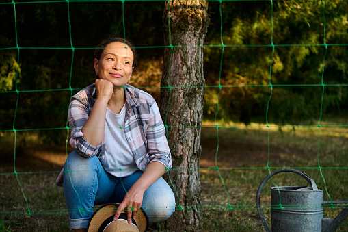 Charming dreamy beautiful Caucasian woman gardener agronomist, cutely smiling while planting seedlings in the backyard of a country house. Growing organic vegetables in eco farm. Agricultural hobby