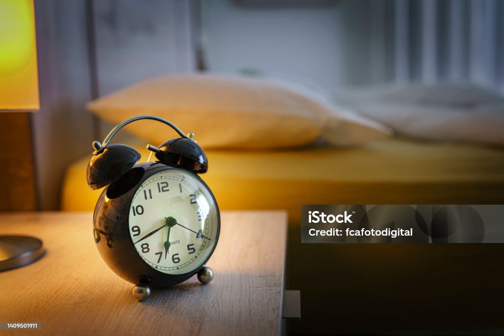 Alarm clock on night table Close up of a black alarm clock at 6:43am on the night table with bed at background. Selective focus on alarm clock. High resolution 42Mp indoors digital capture taken with SONY A7rII and Zeiss Batis 40mm F2.0 CF lens Alarm Clock Stock Photo