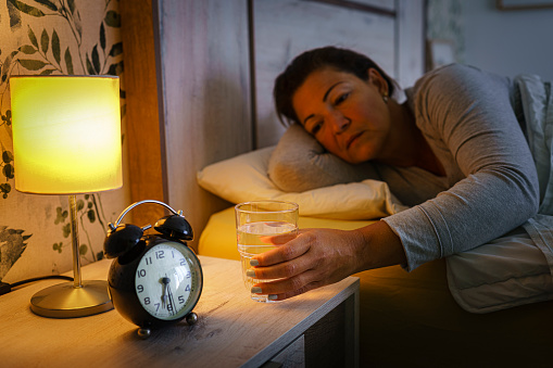 Mature woman lying on bed during the night reaching a glass of water from night table. Selective focus on hand. High resolution 42Mp indoors digital capture taken with SONY A7rII and Zeiss Batis 40mm F2.0 CF lens