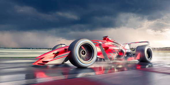 A low angle, close up view of a race car moving at high speed in partially wet conditions with some water spray from wheels and motion blur. The car is on a track under a heavily stormy sky with dark clouds in afternoon / morning.