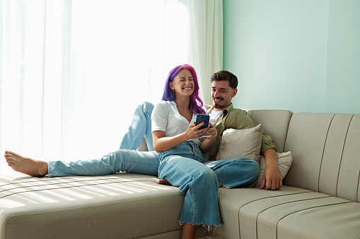 Hipster couple sitting on the couch laughing at memes on the phone at home. Young woman with bright colorful hair and bearded man in the living room having fun. Close up, copy space, background