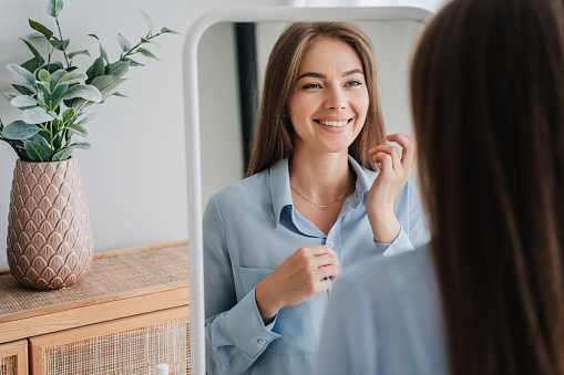 Cheerful young European woman in blue shirt standing at mirror toothy smiling satisfied by her skin health. Successful caucasian blonde woman toothy smiling at home, enjoying sunny morning at bedroom.