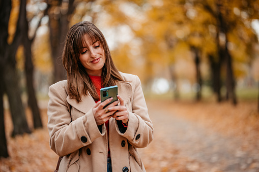 Beautiful young woman walking in the public park during autumn and using smart phone.