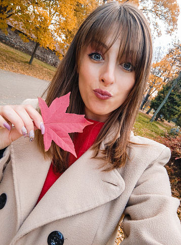 Beautiful young woman holding an autumn leaf and taking a selfie using smart phone in a public park during autumn,