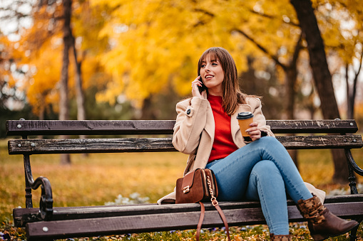 Young woman sitting on the park bench during autumn and having a phone call with a friend. Holding a disposable coffee cup.