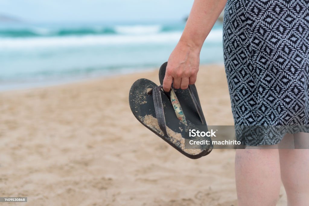 Close-up of a woman in a geometric patterned dress holding sandals as she walks towards the water at the beach. Only Women Stock Photo