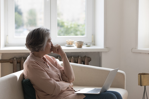 Thoughtful frustrated mature 60s woman holding laptop computer, looking away in deep sad thoughts, thinking over bad concerning news, health problems, sitting on home couch, feeling worried