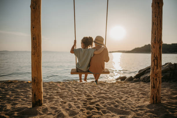 Brother and sister swinging by the sea Little brother and sister sitting on a swing and swinging together on the beach by the sea in sunset. halkidiki beach stock pictures, royalty-free photos & images