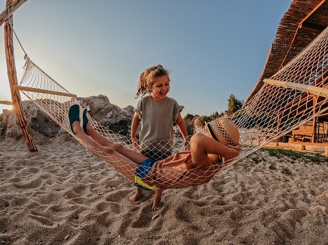 Brother is lying in a hammock, while his little sister is standing next to him, they are having fun together on the beach in sunset.