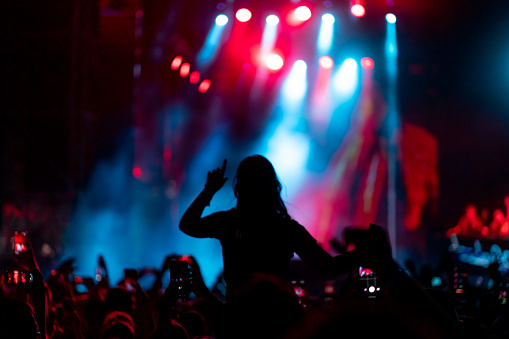 Rear view of a large group of people in front of a music festival stage. Crowd is excited and dancing, raising hands, clapping, punching the air, filming with mobile phones, etc... under blue and red stage lights.