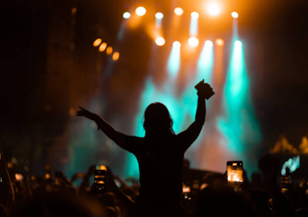 Girl sitting on shoulders at music festival crowd Rear view of a large group of people in front of a music festival stage. Crowd is excited and dancing, raising hands, clapping, punching the air, filming with mobile phones, etc... under orange and teal stage lights. exit festival stock pictures, royalty-free photos & images