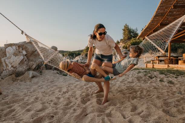 Brother and sister is lying in a hammock, while their mother is swinging them, they are having fun together on the beach in sunset.