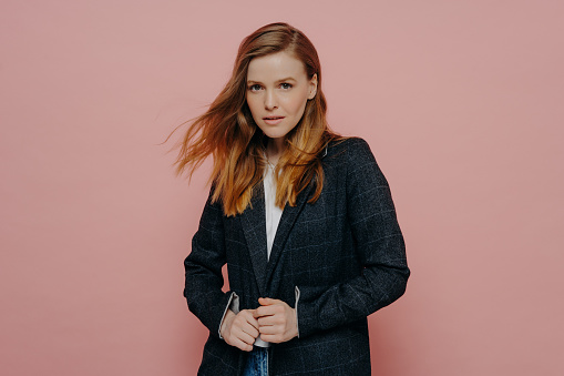 Medium shot of attractive woman with ginger hair looking at camera wearing dark formal jacket and white top posing isolated over pink studio background. Young people concept