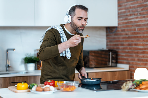 Shot of handsome mature man tasting the food he is preparing while listening music with headphones in the kitchen at home.