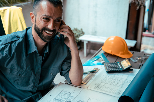 Bearded Male architect talking on the cellphone while working on his blueprint new estate drawing. Male building contractor contracts a new job using phone