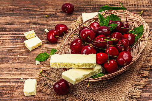 Ripe sweet cherries with fresh mint leaves and white chocolate, traditional summer fruits. Trendy hard light, dark shadow, vintage napkin. Wooden boards background, copy space