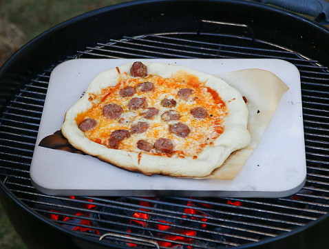 A homemade sausage pizza on a pizza stone over a charcoal grill
