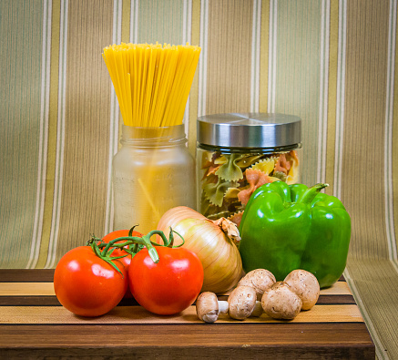 A studio still life featuring an onion, green pepper, fresh mushrooms and three red tomatoes on a wooden cutting board with spaghetti and bow tie pasta in jars.
