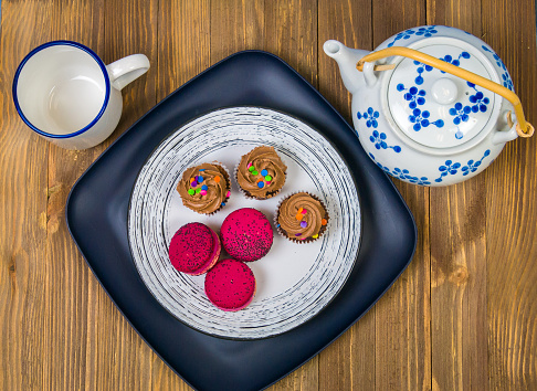 A still life of three chocolate cupcakes and three macarons on a plate with a tea pot and cup in a wooden table.