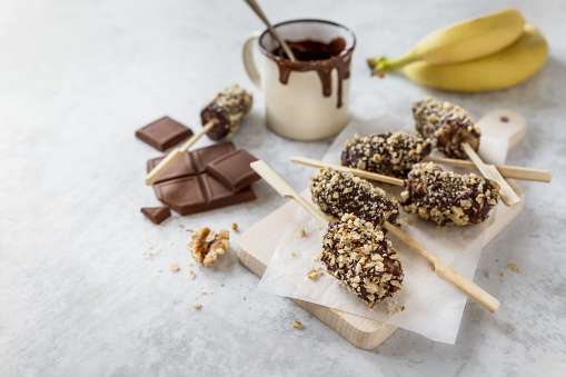 Frozen banana popsicles dipped in melted chocolate and roasted walnuts on wooden board, fresh fruits on skewers easy homemade dessert for kids