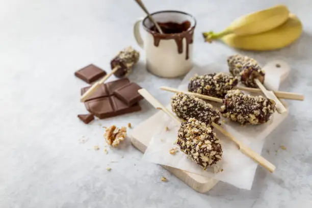 Photo of Frozen bananas on skewers covered with chocolate and nuts on wooden board