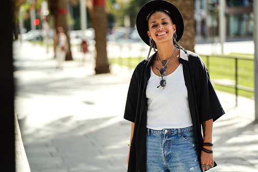 Stylish young girl in fashionable clothes and black hat smiling to the camera, posing on the city street. Real people lifestyle.