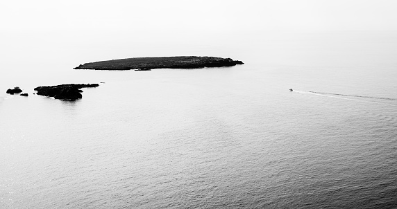 a small boat approaches a group of islets in a huge ocean, Menorca, Balearic Islands, Spain, panoramic view, black and white