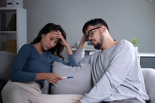 Depressed young Asian woman feeling sad while looking at the positive result of a pregnancy test because husband is not ready to have a child. Young couple feeling sad and disappointed looking at the negative pregnancy test result. Relationship problems concept
