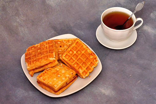 A plate with several fresh Viennese waffles and a cup of hot tea on a saucer with a spoon on a gray abstract background. Close-up.